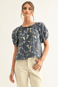 A woman modeling a short-sleeve blouse with puff sleeves, a keyhole neckline, and an abstract blue and cream pattern, paired with cream pants against a plain light background.
