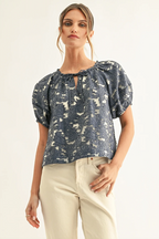 A woman modeling a short-sleeve blouse with puff sleeves, a keyhole neckline, and an abstract blue and cream pattern, paired with cream pants against a plain light background.