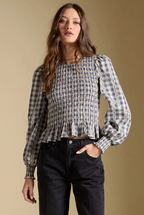 A woman modeling a long-sleeve smocked blouse with a blue, beige, and white gingham pattern, paired with black jeans, against a solid beige background.