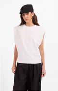 A model wearing a sleeveless white top, black wide-leg pants, a black flat cap, and gold hoop earrings, photographed against a plain white studio background.