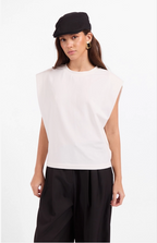A model wearing a sleeveless white top, black wide-leg pants, a black flat cap, and gold hoop earrings, photographed against a plain white studio background.