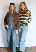 Two women posing together in front of a white wall, wearing vintage-style clothing. The woman on the left wears a brown zip-up jacket over a gray top with blue jeans, and the woman on the right wears a green and cream striped knit polo sweater with blue jeans.