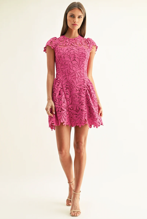 A woman modeling a bright pink floral lace mini dress with cap sleeves and a flared skirt, paired with nude high-heeled sandals, against a plain off-white studio background.