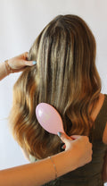 A woman with long, wavy brown and blonde highlighted hair is shown from behind while being brushed with a light pink hairbrush. Her hands are visible holding her hair and the brush.