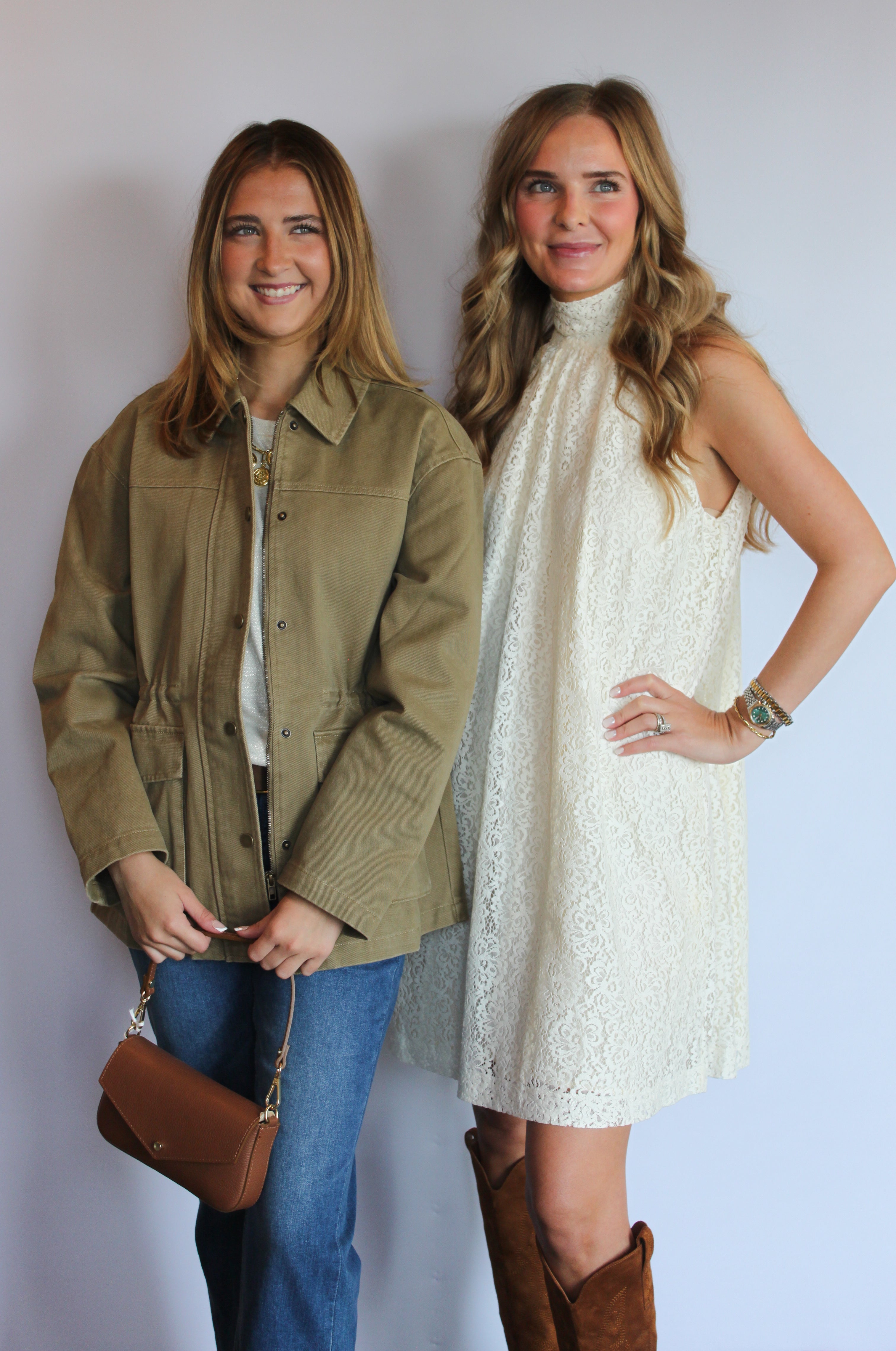 Two women posing together against a light gray studio background. The woman on the left wears an olive green utility jacket over a white top and blue jeans, holding a brown leather handbag.
