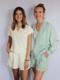 Two women standing side-by-side wearing matching light-colored loungewear sets. The woman on the left wears a cream short-sleeve top and shorts set, while the woman on the right wears a mint green zip-up hoodie and matching shorts.