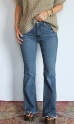 A person wearing medium-wash blue flare jeans with a frayed hem, styled with a beige knit top and brown platform sandals, standing against a white wall on a patterned rug.