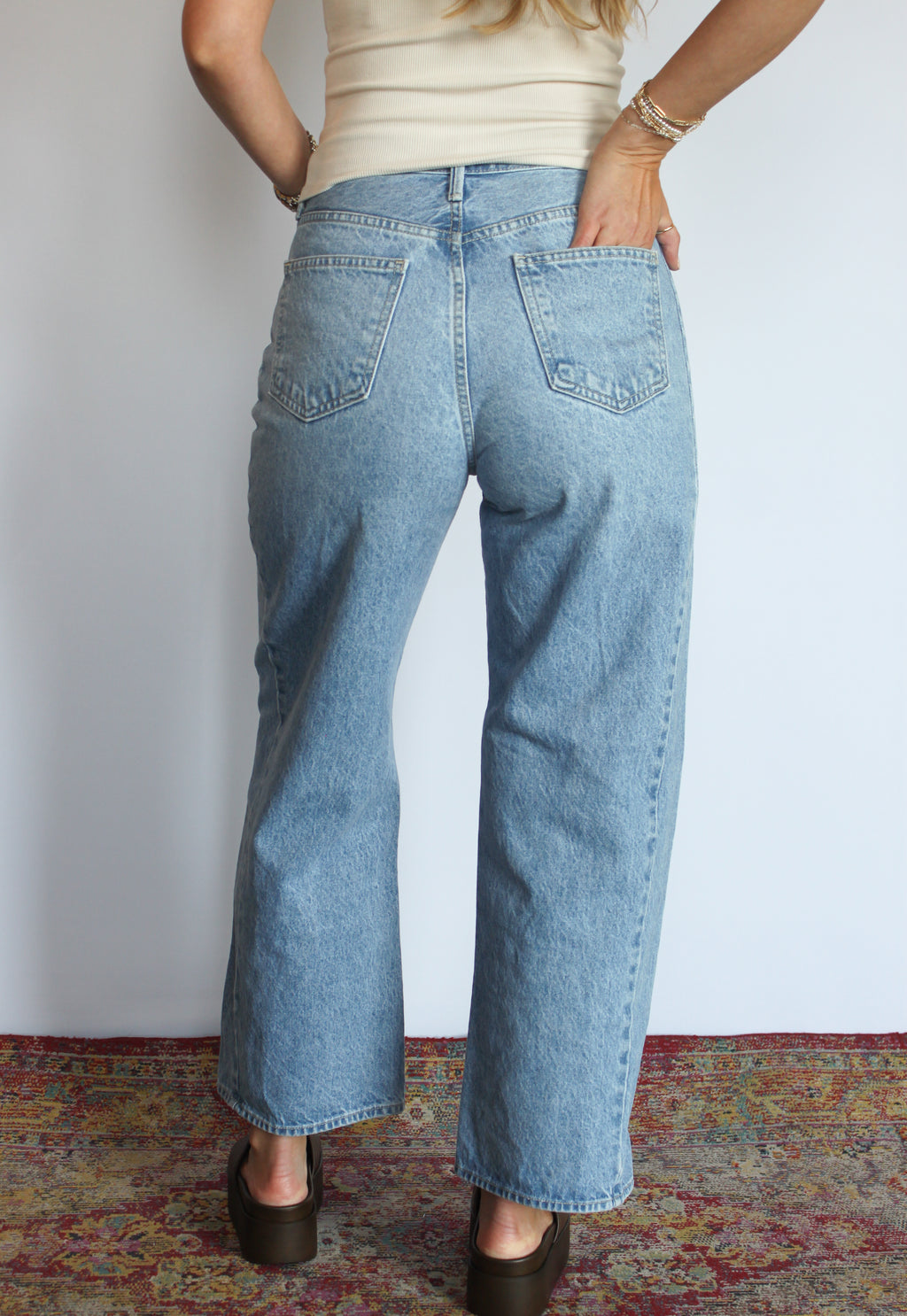 A person shown from the back wearing light-wash blue wide-leg jeans, a cream ribbed top, and dark brown shoes, standing against a plain white wall with a patterned rug visible at the bottom.