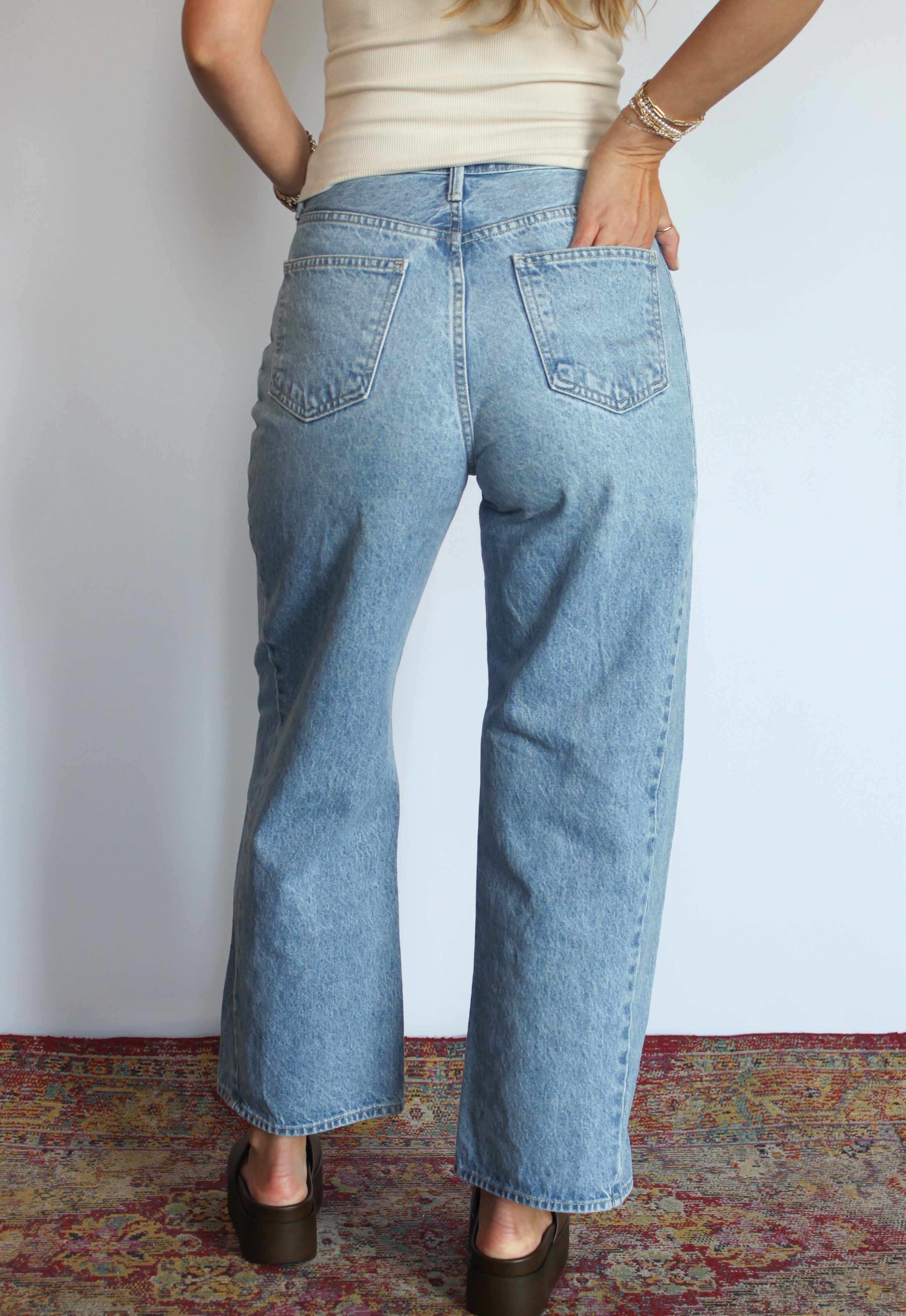 A person shown from the back wearing light-wash blue wide-leg jeans, a cream ribbed top, and dark brown shoes, standing against a plain white wall with a patterned rug visible at the bottom.