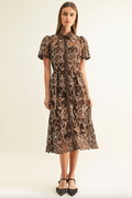 A woman models a brown short-sleeve midi dress with an intricate floral lace overlay and a button-up front, paired with black pointed-toe heels against a plain off-white studio background.
