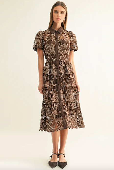 A woman models a brown short-sleeve midi dress with an intricate floral lace overlay and a button-up front, paired with black pointed-toe heels against a plain off-white studio background.