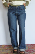 A person wearing a pair of straight-leg medium wash blue jeans, styled with a green and cream striped sweater and brown platform sandals, standing on a patterned rug against a white wall.