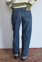 A person shown from the back wearing blue straight-leg denim jeans with cuffed hems, paired with an olive green and cream sweater, standing on a patterned rug against a plain white wall.