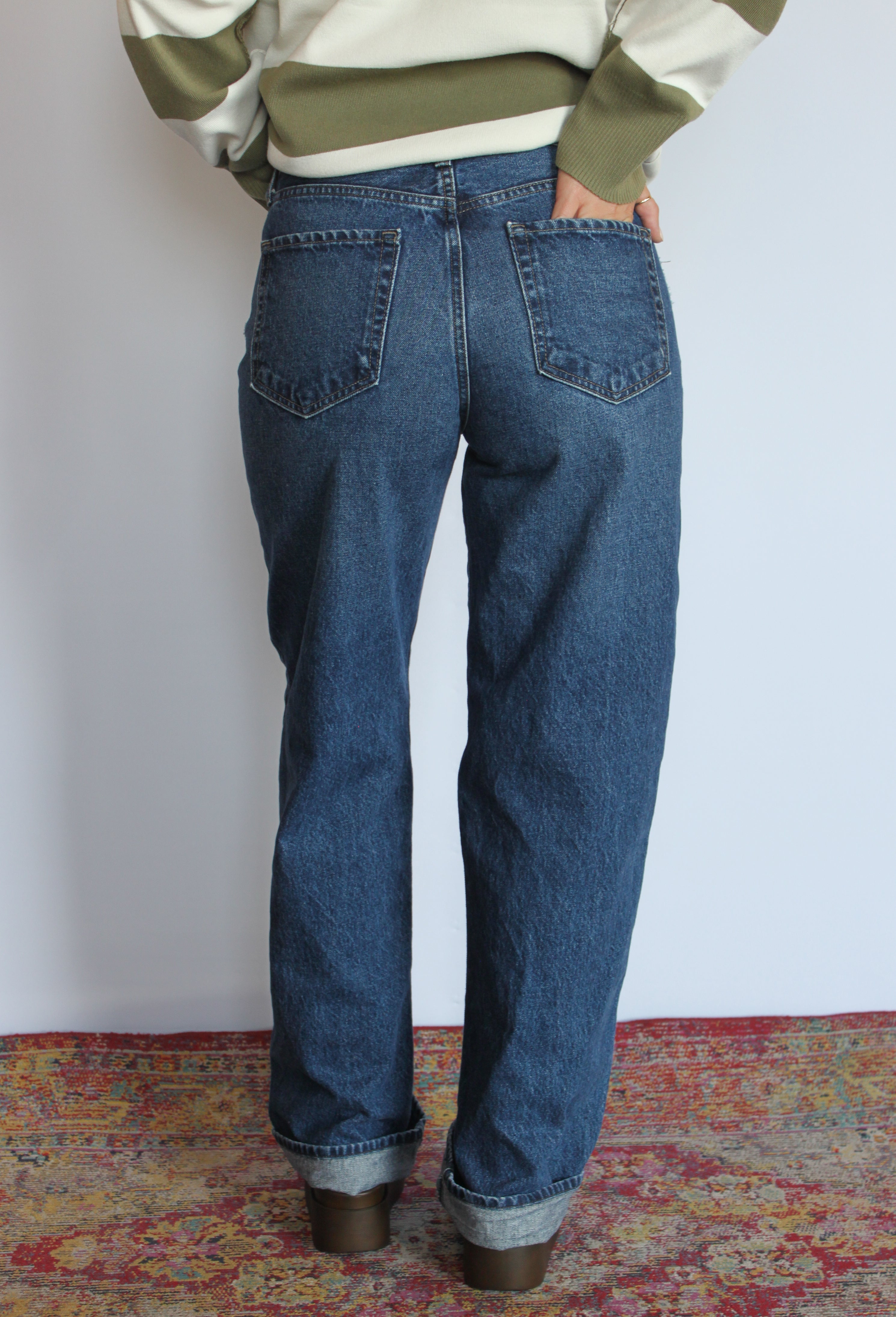 A person shown from the back wearing blue straight-leg denim jeans with cuffed hems, paired with an olive green and cream sweater, standing on a patterned rug against a plain white wall.