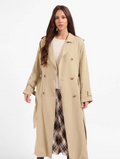 A woman modeling a long beige trench coat over a cream top and a plaid skirt, photographed against a plain white studio background.