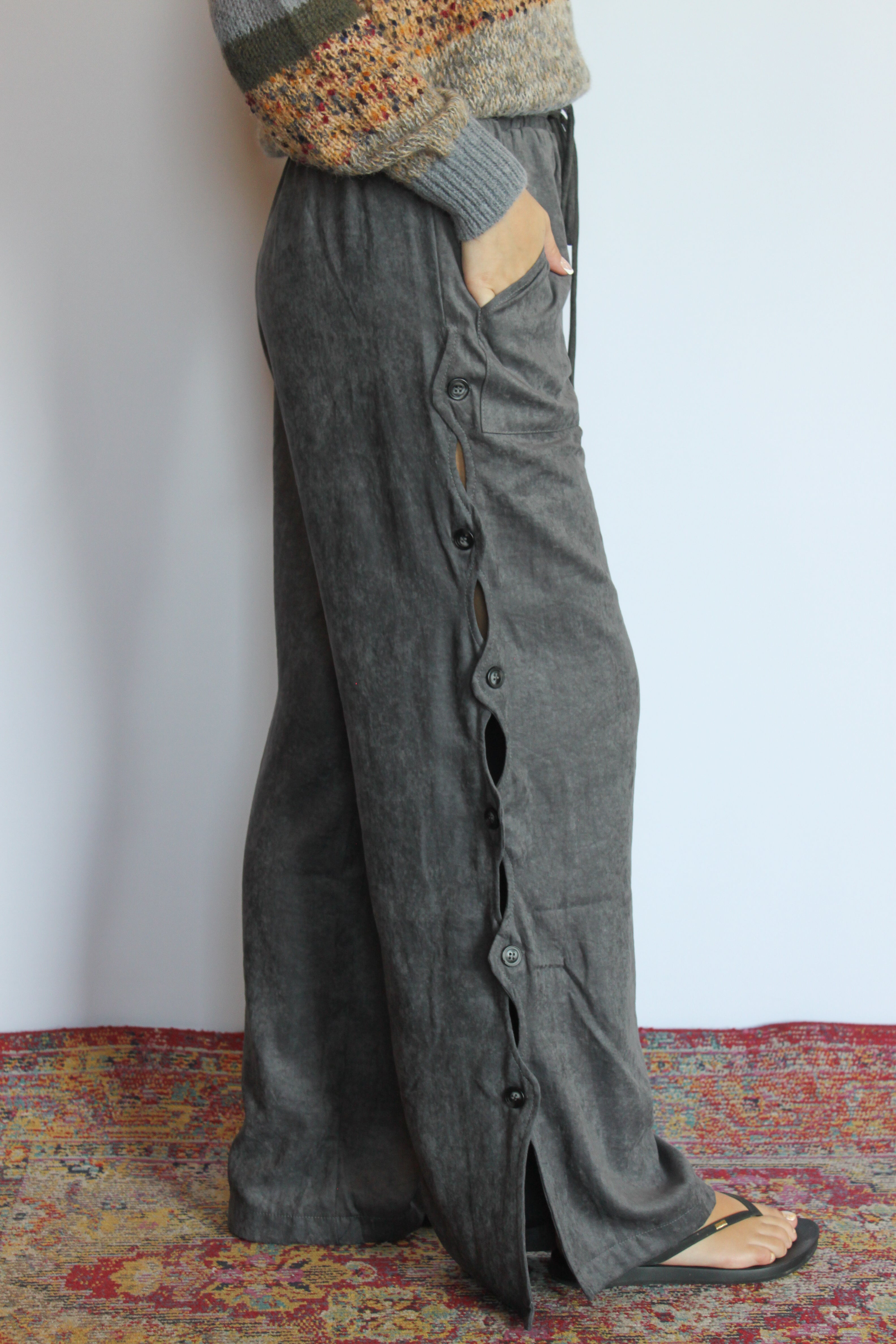 A person wearing loose, wide-leg gray linen pants with side buttons and a drawstring waist, standing on a patterned rug against a plain white wall.