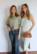 Two women posing together against a plain white wall. The woman on the left wears a cropped sage green button-up shirt with blue jeans, and the woman on the right wears a light gray sleeveless midi dress with a brown leather clutch.