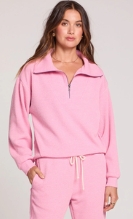 A woman modeling a light pink two-piece tracksuit featuring a quarter-zip sweatshirt and matching sweatpants with a drawstring waist, set against a plain light gray studio background.