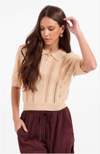 A woman modeling a beige short-sleeve knit polo sweater with eyelet and lace-up front details, paired with dark brown drawstring pants, against a plain white studio background.