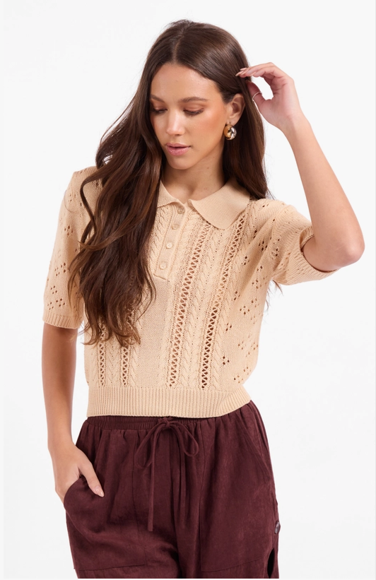 A woman modeling a beige short-sleeve knit polo sweater with eyelet and lace-up front details, paired with dark brown drawstring pants, against a plain white studio background.