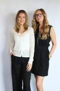 Two women standing side-by-side against a plain light gray wall. The woman on the left wears a white textured long-sleeve top over a white camisole, paired with black pants.