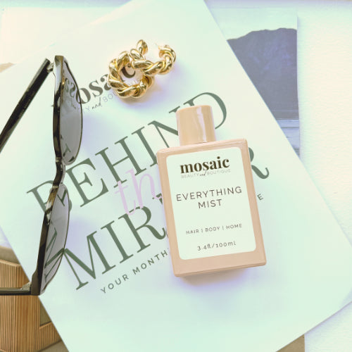 A bottle of mosaic Everything Mist hair, body, and home fragrance in a pale peach bottle with a white label, styled on top of a magazine page with sunglasses and a gold chain.