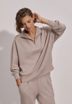 A woman modeling a light taupe half-zip pullover sweatshirt and matching sweatpants against a plain light gray studio background.