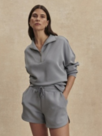 A woman modeling a light gray matching lounge set consisting of a half-zip long-sleeve top and coordinating shorts, set against a textured beige background.