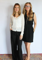Two women standing side-by-side in an indoor setting against a white wall, wearing casual outfits. The woman on the left wears a white long-sleeve lace top with black wide-leg pants, and the woman on the right wears a black sleeveless mini dress with glasses.