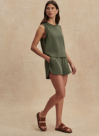 A woman modeling a sleeveless olive green top and matching shorts set, paired with brown slide sandals, against a textured beige studio backdrop.