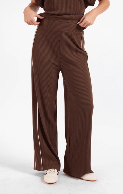 A person wearing brown ribbed wide-leg pants with thin light beige side stripes, paired with a matching brown ribbed top and light-colored sneakers, photographed against a plain light gray studio background.