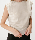 A woman wearing a sleeveless, crew-neck top in a light beige or cream color, paired with black pants, against a plain white background.