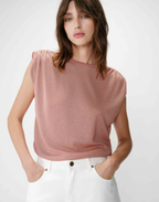 A woman wearing a sleeveless dusty rose knit top and white pants, standing against a light gray studio background.